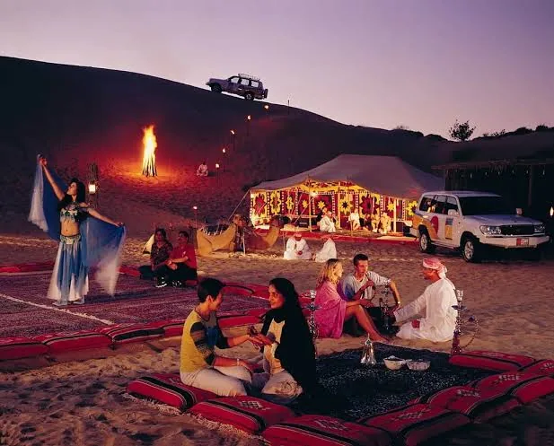 Guests seated on traditional cushions with an illuminated tent and dancer at a Qatar desert camp in the evening