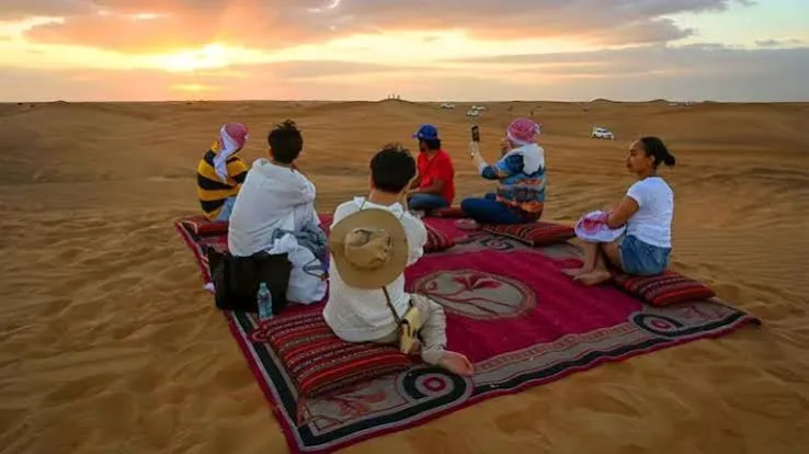 Guests seated on a red majlis rug on a sand dune watching the sunset on a Qatar desert safari and camp