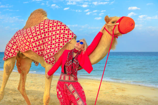 Happy blonde woman with camel on a sand beach of Khor al Udaid in Persian Gulf of southern Qatar. Caucasian tourist after camel ride, a popular tour in Middle East of Arabian Peninsula. Sunn blue sky.