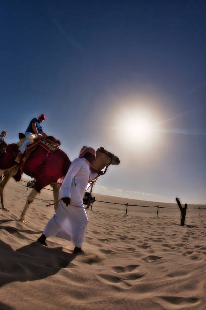 Doha, Qatar - January 28 2010 : tourists are having fun as riding camel in the desert with a man with head scarf and thaw helping